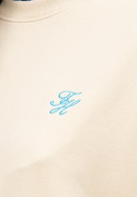 Ύφασμα σε κρεμ απόχρωση με ένα μικρό, ανοιχτά γαλάζιο κεντημένο μονόγραμμα που απεικονίζει διαπλεγμένα γράμματα "F" και "H".