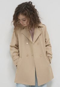 Beige doppelt-knöpfter Mantel mit großen Knöpfen, breiten Revers und Seitentaschen, getragen über einem hellrosa Oberteil. Strukturierter, weicher Stoff.