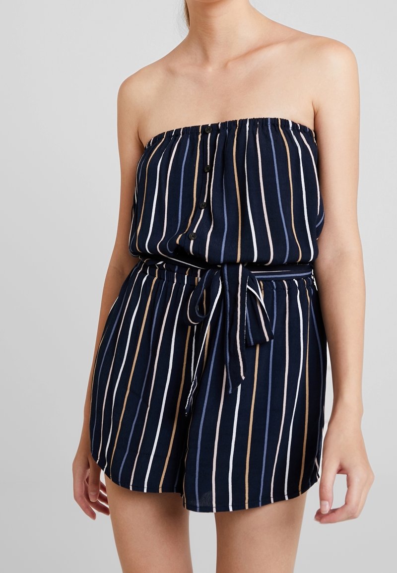 Beżowo-białe pionowe paski w granatowym kombinezonie bez ramiączek, z elastycznym pasem w talii i szczegółem związania paska. Guziki z przodu.