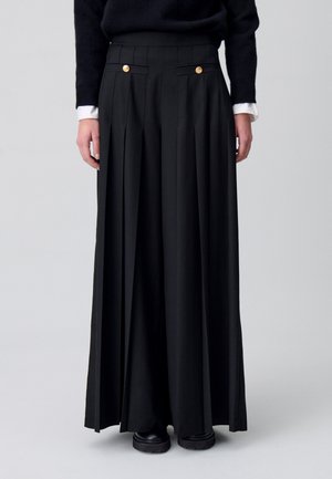 Pantalons larges plissés noirs avec accents de boutons dorés, taille haute, design ajusté et texture lisse. S'ouvre au niveau de la cheville.