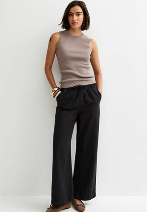 Kobieta ubrana w bezrękawnikowy taupe prążkowany top, czarne szerokie spodnie, brązowe baleriny i bransoletki, stojąca z rękami w kieszeniach na tle białej ściany.