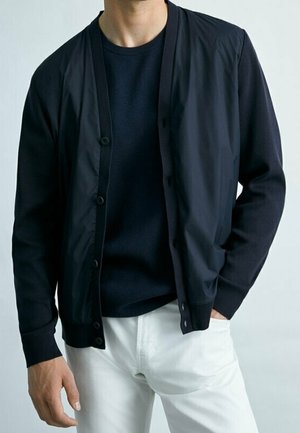 Homme portant un cardigan boutonné bleu marine foncé sur une chemise bleu marine et un pantalon blanc, debout avec une main dans la poche.