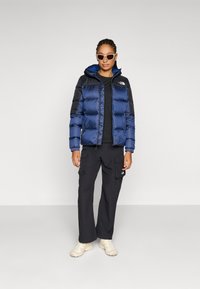 Blauwe pufferjack met zwarte accenten, ritssluiting aan de voorkant, capuchon en logo. Gecombineerd met zwarte cargobroek en lichtgekleurde schoenen. Witte zonnebril.