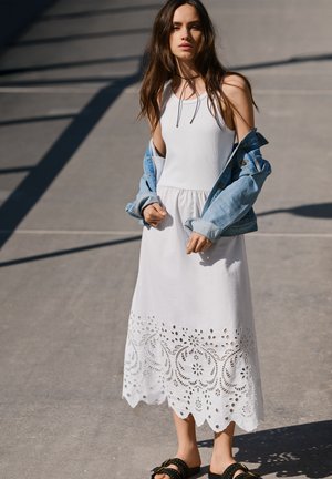 Jeune femme portant une robe blanche sans manches avec un ourlet en dentelle œillet, une veste en jean claire tombant des épaules, et des sandales noires cloutées en extérieur.