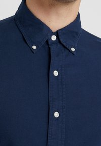 Námorně modrá košile na knoflíky s klasickým límcem, se bílými knoflíky a strukturovanou tkaninou. Detailní pohled na výstřih a přední zapínání.