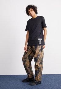 Μαύρο βαμβακερό μπλουζάκι με λευκό λογότυπο Adidas στο στρίφωμα, συνδυασμένο με φαρδιά παντελόνια με σχέδιο tie-dye σε γήινες αποχρώσεις και μαύρα αθλητικά παπούτσια.