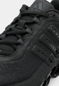 Detailní záběr černé běžecké boty Adidas s texturovaným síťovaným svrškem, reflexními detaily na tkaničkách a podrážkou ve tvaru pružiny.