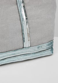 Coin d'un sac en tissu gris avec un galon vertical et horizontal de sequins argentés sur une surface blanche.