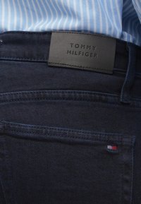 Mørkeblå jeans med en glatt tekstur, som har en svart læretikett merket "TOMMY HILFIGER" og en liten rød, hvit og blå logo på lommen.