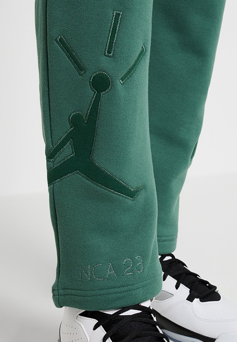 Des joggings en polaire verte présentent une silhouette en relief d'un joueur sautant pour un ballon de basket, avec des détails de couture portant l'inscription "NCA 23." Accentués par des baskets noires.