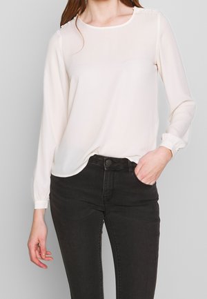 Blouse à manches longues couleur crème avec un col arrondi, de délicats boutons décoratifs aux épaules et un tissu fluide, associée à un jean noir.