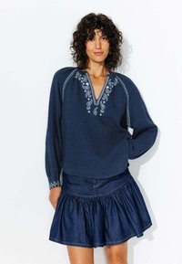 Blouse brodée bleu marine à manches longues et décolleté en V, associée à une jupe en denim avec un ourlet froncé.