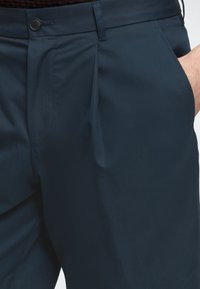 Pantalon bleu marine avec pli à l'avant, poches latérales, passants de ceinture et fermeture par bouton, porté avec une main dans la poche.