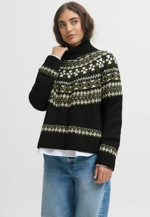Femme portant un pull à col roulé noir et vert à motifs avec un jean bleu ample, posant avec une main touchant son cou.