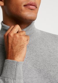 Pull col roulé gris en maille douce et texturée avec un col et des poignets côtelés, mettant en avant un gros plan d'une main ajustant le col.