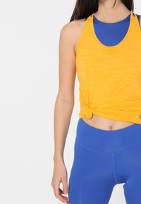 Gul sportstopp med knuten design, lager över ett blått baslager, ihop med blå leggings, slät textur, figurnära passform.