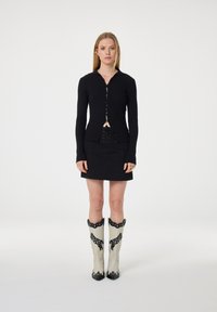 Vrouw in een zwarte geribbelde knoptruien, zwarte mini-jurk met vetersluitingen en beige cowboylaarzen met zwarte patronen, rechtop staand.