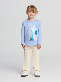 Jasnoniebieska koszula z długim rękawem z błyszczącym nadrukiem postaci, zestawiona z kremowymi, prążkowanymi spodniami o rozkloszowanym kroju i białymi tenisówkami.