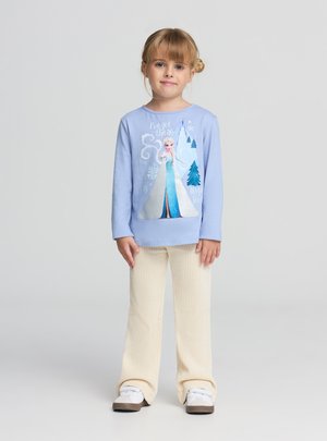 Jasnoniebieska koszula z długim rękawem z błyszczącym nadrukiem postaci, zestawiona z kremowymi, prążkowanymi spodniami o rozkloszowanym kroju i białymi tenisówkami.