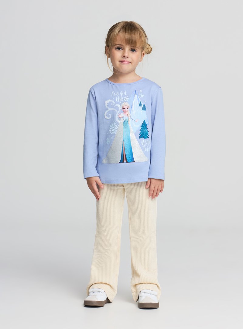 Jasnoniebieska koszula z długim rękawem z błyszczącym nadrukiem postaci, zestawiona z kremowymi, prążkowanymi spodniami o rozkloszowanym kroju i białymi tenisówkami.