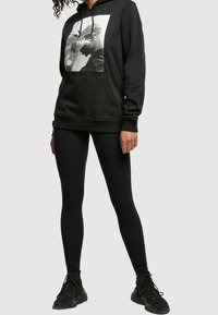 Osoba v černé mikině s grafikou Tupaca, černých legínách a černých teniskách, stojící na pozadí s jednoduchým světlým pozadím.