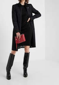 Cappotto lungo nero su un abito nero aderente, con stivali al ginocchio neri lucidi e una piccola borsa rossa con manico e hardware argentato.