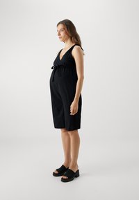 Zwart zwangerschapsjurk met een V-hals, strikdetail in de taille, knie-lengte, zachte stoftextuur. Gecombineerd met zwarte open-hak blokhakken.