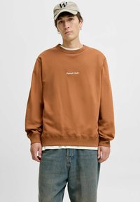Felpa marrone in misto cotone, con polsini e orlo a costine, logo ricamato in bianco, indossata sopra una maglietta bianca e jeans chiari.