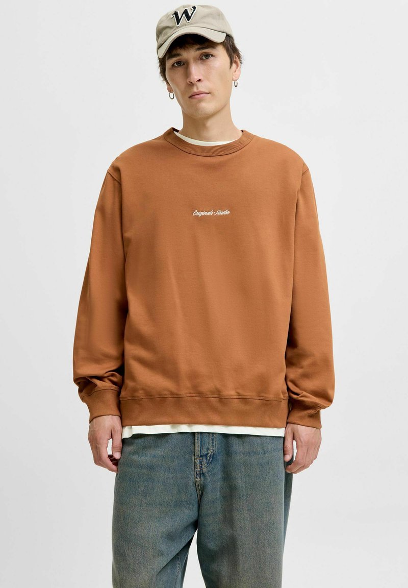Felpa marrone in misto cotone, con polsini e orlo a costine, logo ricamato in bianco, indossata sopra una maglietta bianca e jeans chiari.