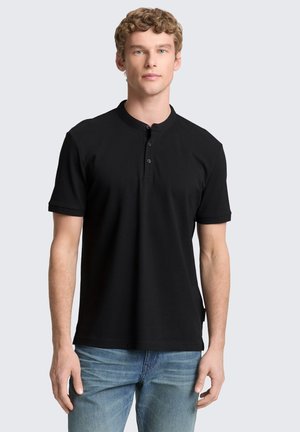 Młody mężczyzna z kręconymi włosami, ubrany w czarną koszulkę henley z krótkim rękawem i niebieskie dżinsy, stoi na tle jednolitego jasnego tła.