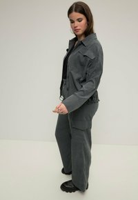Szara kurtka typu utility z cordurowego materiału, o luźnym kroju, z dużymi kieszeniami i ściągniętą talią, zestawiona z dopasowanymi spodniami typu wide-leg i czarnymi botkami.