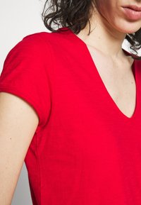 T-shirt di cotone rosso con scollo a V, maniche corte e una texture morbida. Il tessuto appare leggermente stropicciato, esaltando un look informale.