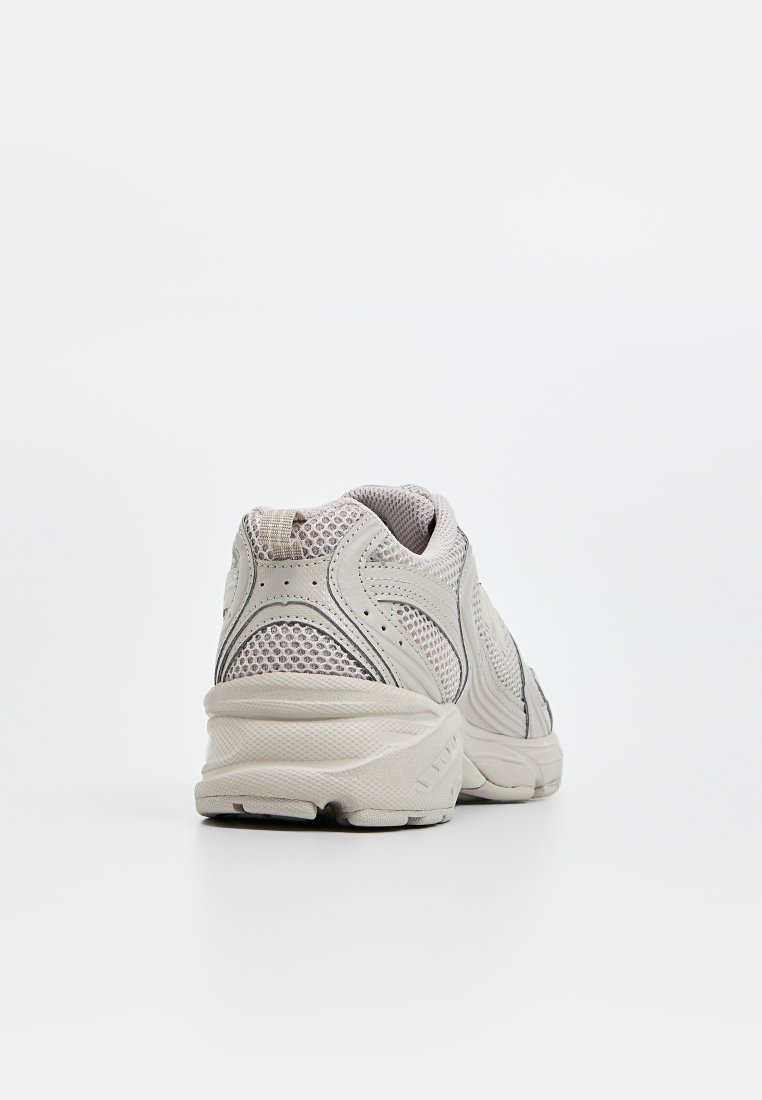 Zapatillas deportivas de color gris claro con parte superior de malla, cuello acolchado y suela texturizada. Presenta costuras sutiles y una lengüeta de goma en la parte trasera.