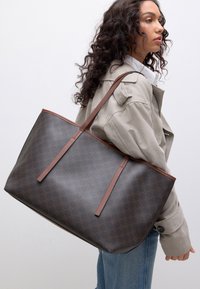 Sac fourre-tout marron à motif avec une texture lisse, des anses marron, et un design rectangulaire spacieux, porté sur l'épaule par une personne.