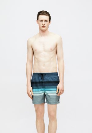 Junger Mann mit nassen Haaren, der gestreifte blaue Badeshorts trägt, steht vor einem schlichten hellgrauen Hintergrund und schaut nach vorne.