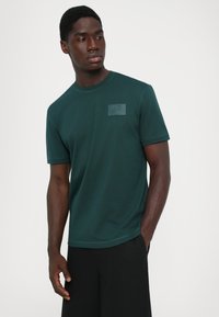 Tumši zaļš kokvilnas T-krekls ar īsām piedurknēm un apaļu kaklu. Augšējā kreisajā krūtīs redzama reljefa logotipa uzlīme. Gluda tekstūra.