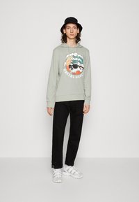 Felpa verde chiaro con stampa grafica, pantaloni neri, sneakers bianche e un cappello a secchiello nero. La felpa presenta un design con onde e teschi.