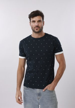 Bărbat cu păr închis la culoare și barbă, purtând un tricou negru cu mici modele albe de ancore și blugi deschiși la culoare, stând cu o mână în buzunar.