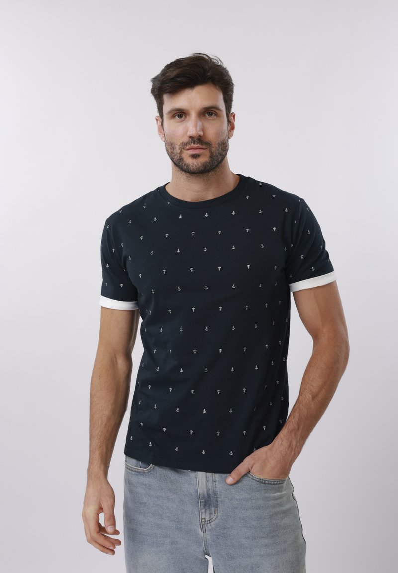 Άνδρας με σκούρα μαλλιά και γενειάδα, που φοράει μαύρο t-shirt με μικρά λευκά μοτίβα άγκυρας και ανοιχτόχρωμο τζιν, στέκεται με το ένα χέρι στην τσέπη.