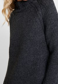 Tlustý tmavě šedý pletený svetr s vysokým límcem, s žebrovanou texturou a volným střihem, zobrazující detailní šití podél rukávů.