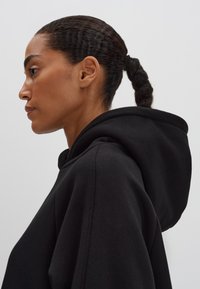 Felpa nera con cappuccio realizzata in un tessuto morbido. Presenta una vestibilità ampia, un cappuccio arrotondato e cuciture visibili lungo i bordi. Vista laterale.