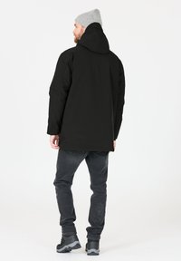 Musta vedenpitävä parka, jossa on huppu, suora leikkaus ja pitkät hihat. Yhdistetty tummanharmaisiin farkkuihin ja kestäviin mustiin saappaisiin.