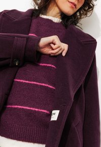 Manteau en laine violet avec un large col, poignets boutonnés, porté sur un pull bordeaux avec des rayures roses. Texture douce, design structuré.