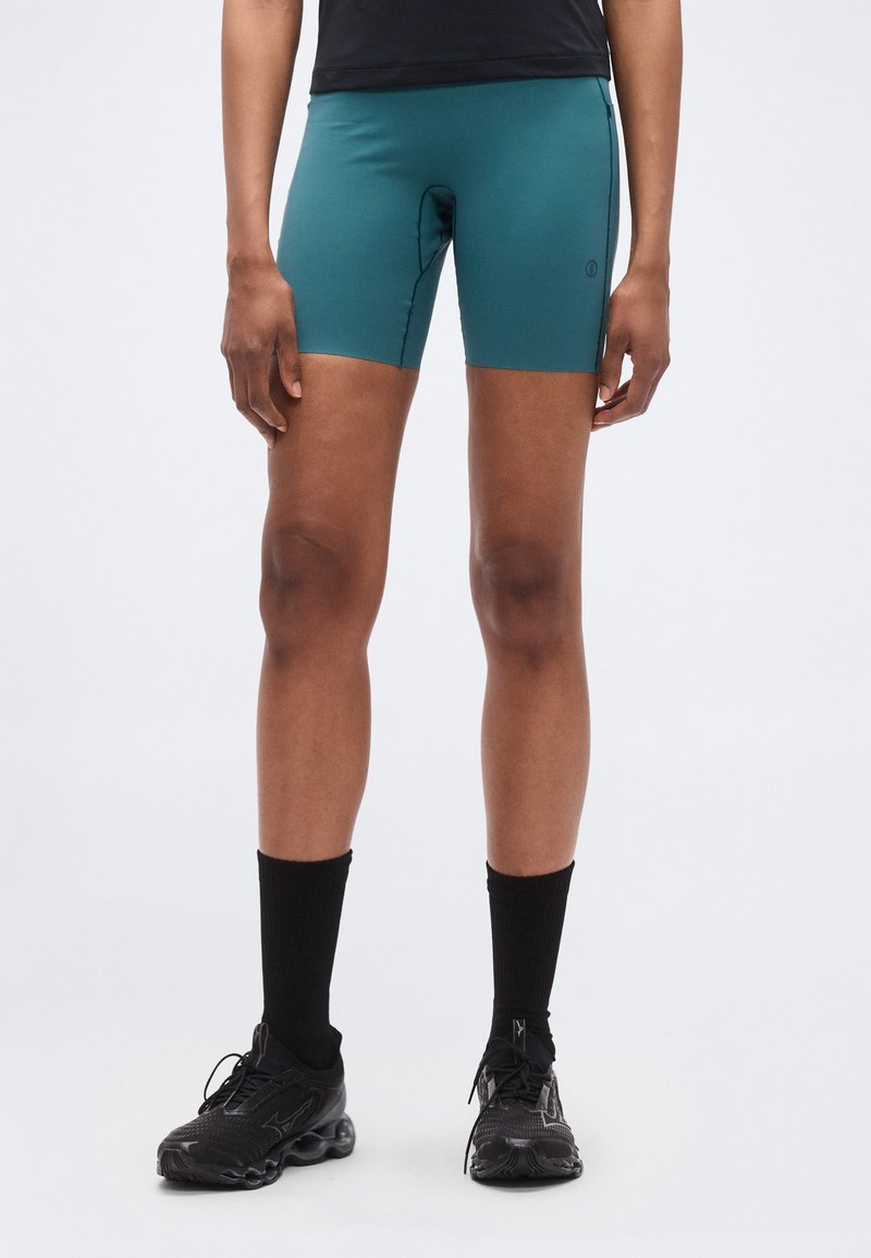 Τιρκουάζ αθλητικό σορτς από ελαστικό ύφασμα, με εφαρμοστή σχεδίαση και μήκος μέχρι το μέσο του μηρού. Φοριούνται με μαύρες κάλτσες και αθλητικά παπούτσια.