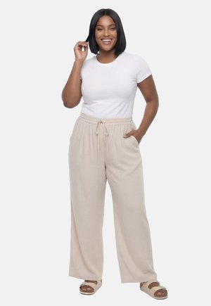 Uśmiechnięta kobieta o czarnych włosach bob, ubrana w białą koszulę z krótkim rękawem, beżowe spodnie o szerokich nogawkach ze sznurkiem i beżowe sandały, stojąca.