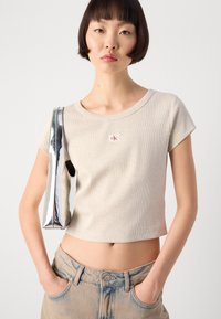 Beige kortermet t-skjorte med ribbet tekstur og et lite rødt logo-patch. Kombinert med falmede, høy midje jeans.