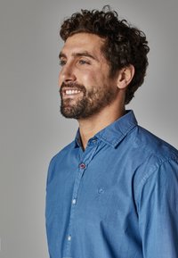 Camisa de botones azul hecha de algodón, con cuello, detalle de botones rojos en contraste y un logo bordado en el pecho.
