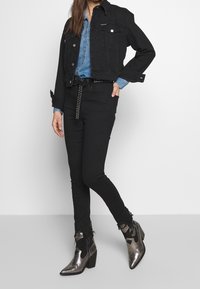 Chaqueta de mezclilla negra, camisa de mezclilla azul clara debajo, jeans negros con cinturón de tachuelas y botines metálicos con punta afilada.