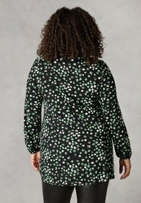 Langärmlige Tunika aus schwarzem Stoff mit kleinen grünen und weißen Blumenmustern, leicht abgerundetem Saum und lockerer Passform.