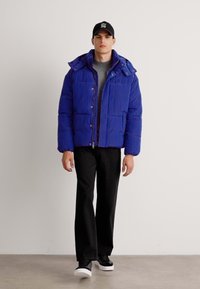 Blå pufferjakke med hette, med en quiltet design og store lommer foran. Kombinert med svarte bukser og hvite sneakers.